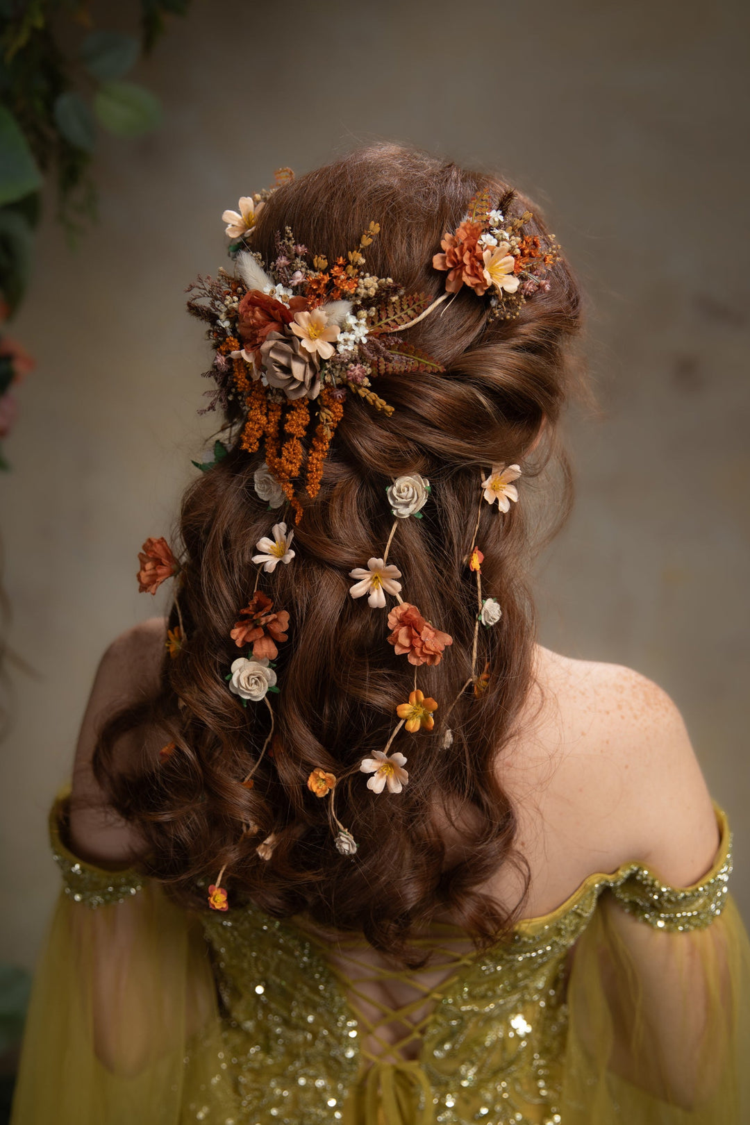 A woman shown from behind wearing a brown half-up hairstyle adorned with a floral hairpiece and dangling dried flowers in orange, cream, and white. She is wearing a off-the-shoulder embellished dress against a soft neutral background.