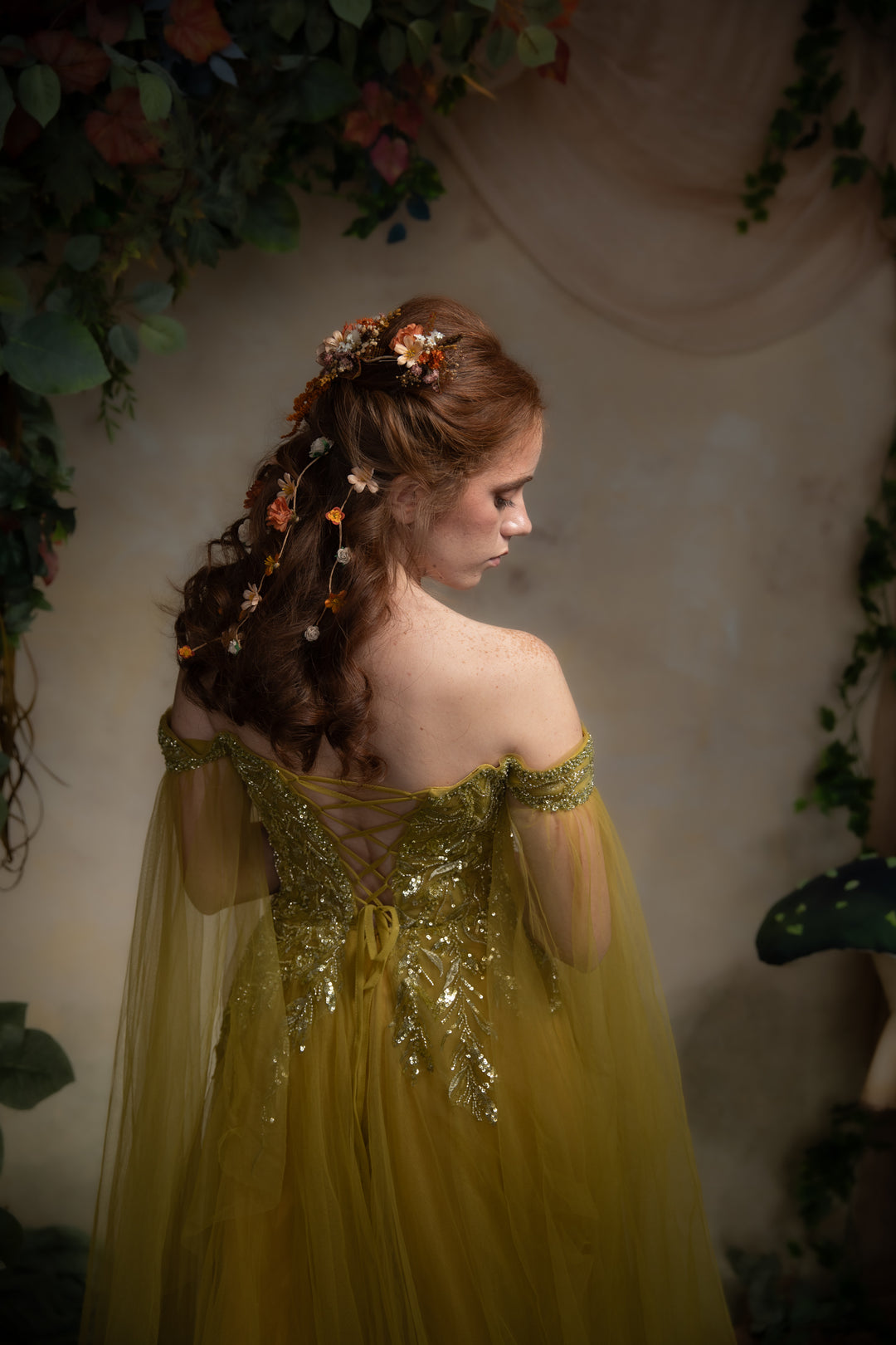 A woman with long auburn hair adorned with flowers, wearing an off-the-shoulder tulle gown with gold embroidery, photographed in a whimsical garden-themed setting.