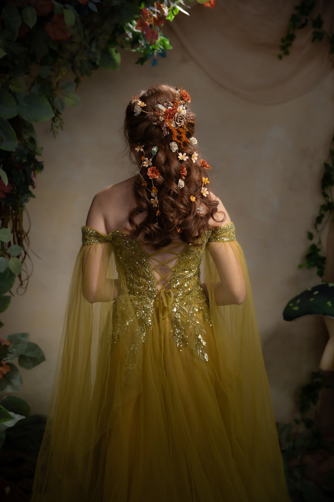 A woman viewed from behind wearing an off-the-shoulder greentulle gown with a heavily embellished gold back and long wavy brown hair decorated with orange and white flowers. She is framed by a soft-focus background of green foliage and a mushroom prop.