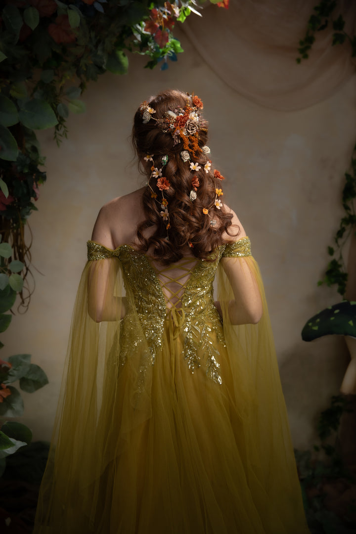 A woman viewed from behind wearing an off-the-shoulder greentulle gown with a heavily embellished gold back and long wavy brown hair decorated with orange and white flowers. She is framed by a soft-focus background of green foliage and a mushroom prop.