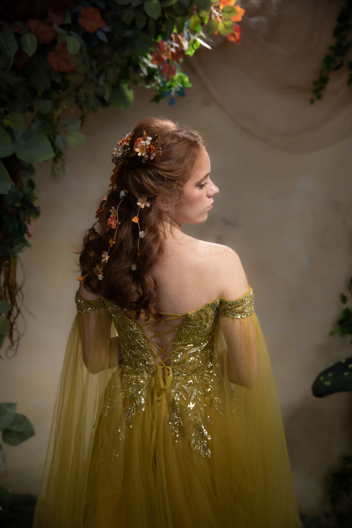 A woman wearing an off-the-shoulder green gown with gold sequined embroidery and sheer sleeves, photographed from the back in a softly lit setting with floral greenery.