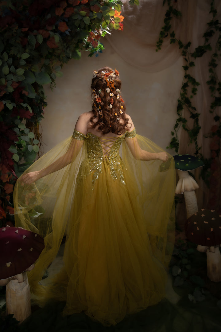 A woman stands with her back to the camera wearing a flowing green tulle gown with gold embellishments and sheer sleeves, posed in a whimsical garden-like setting with flowers, vines, mushrooms, and draped fabric.