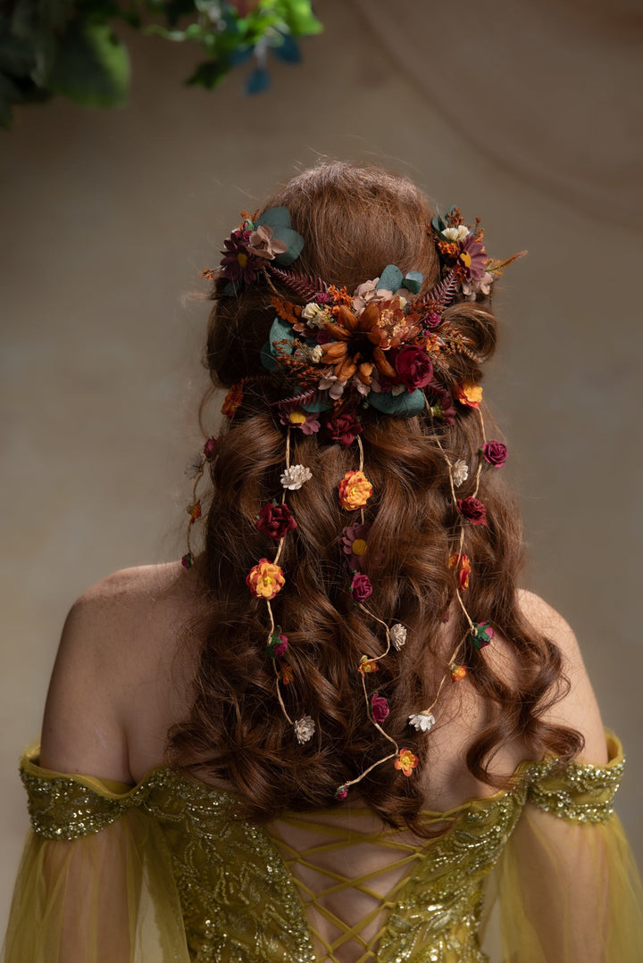 A woman shown from behind wearing an elaborate updo hairstyle adorned with flowers, leaves, and small hanging botanical ornaments. She is wearing a gold off-the-shoulder gown against a soft beige background.