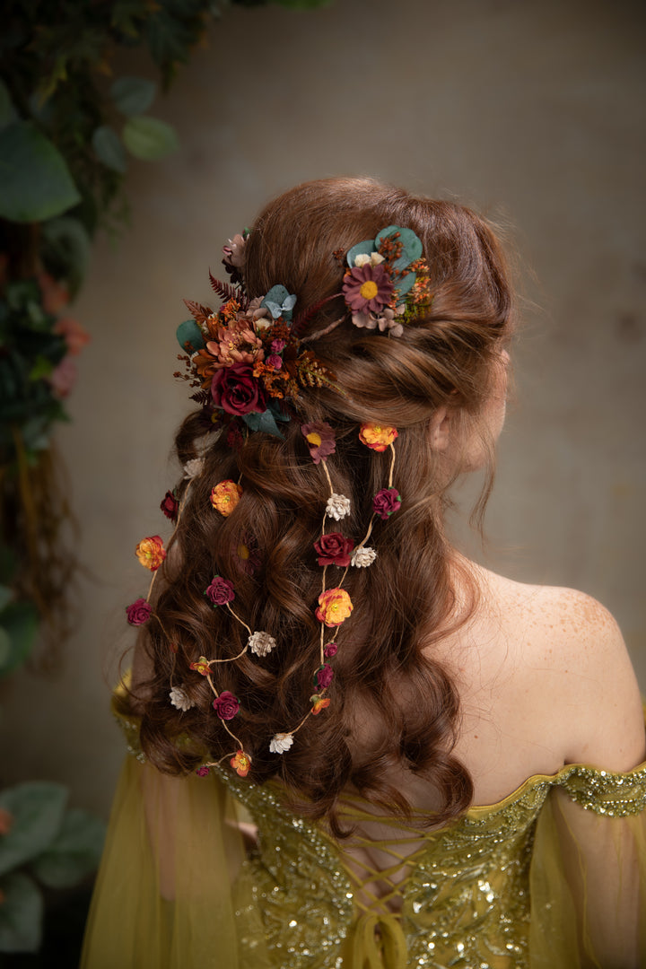 A woman shown from the back wearing a decorative floral hair accessory with flowers and small vine accents in warm autumn colors, paired with a shimmering olive green dress against a softly blurred neutral background.