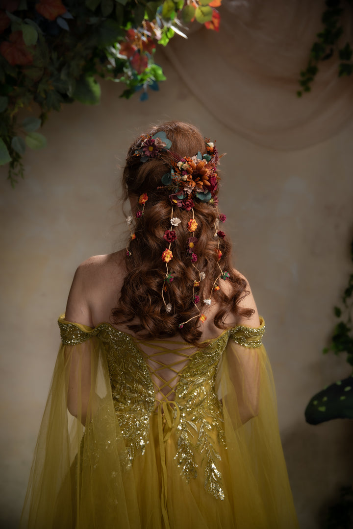 A woman viewed from behind wearing a flowing gown with gold embellishment and a decorative floral hairpiece with dangling orange and burgundy accents, set against a softly lit beige wall with hanging greenery.
