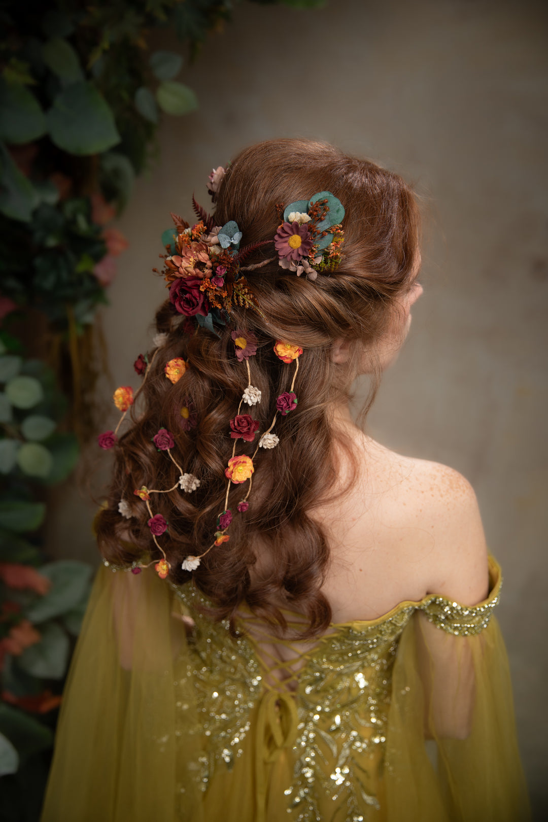 A woman shown from the back wearing a off-the-shoulder dress and a decorative earthy tones hair accessory with flowers and small hanging blossoms, set against a soft gray background with blurred greenery on the left.