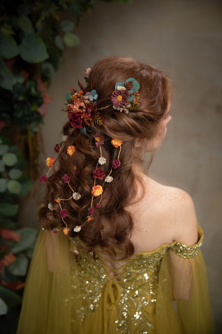 A woman shown from the back wearing a off-the-shoulder dress and a decorative earthy tones hair accessory with flowers and small hanging blossoms, set against a soft gray background with blurred greenery on the left.