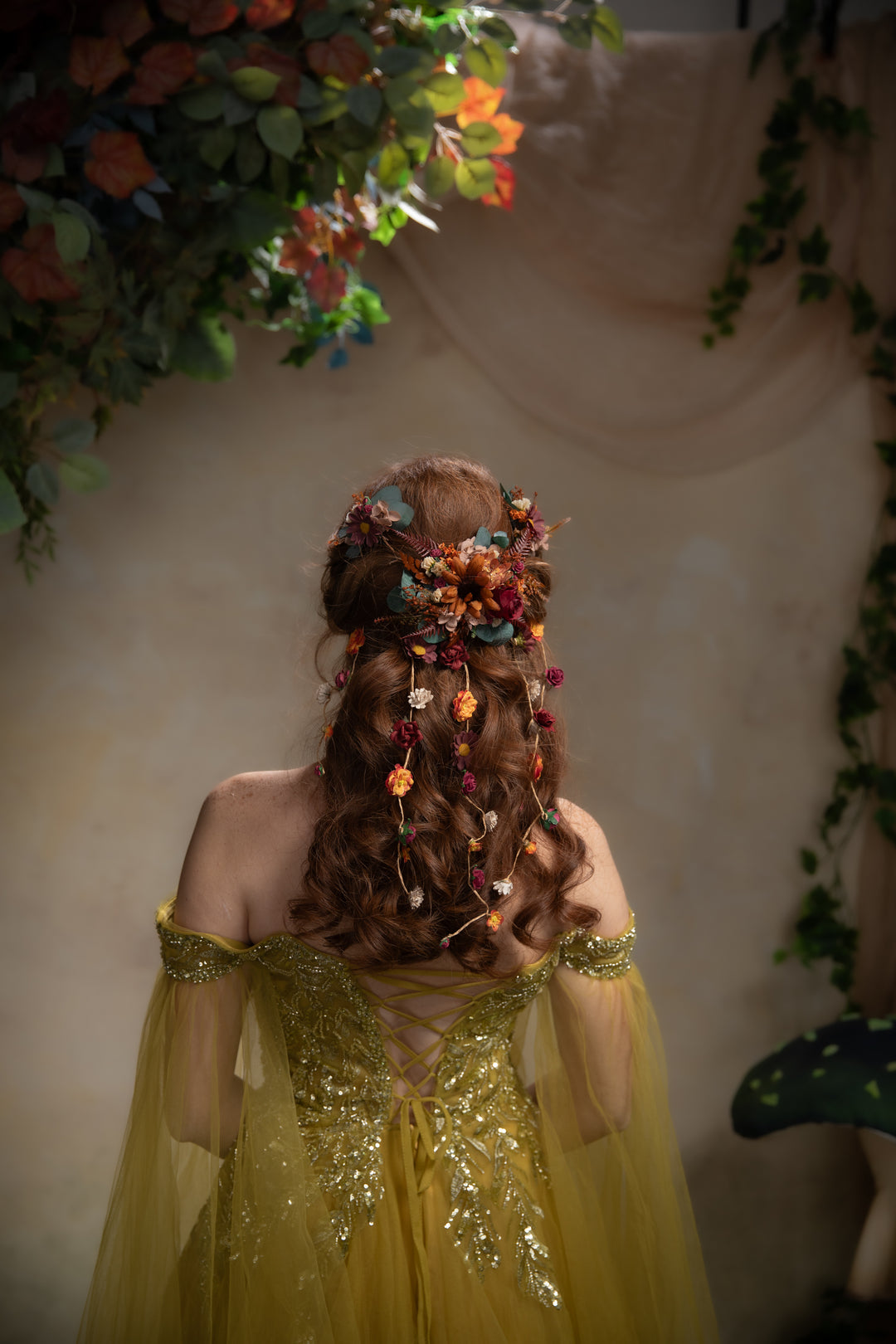 A woman seen from behind wearing a shimmering green off-the-shoulder gown and an elaborate brown hairpiece with colorful floral and leaf accents, posed against a soft beige backdrop with hanging foliage.