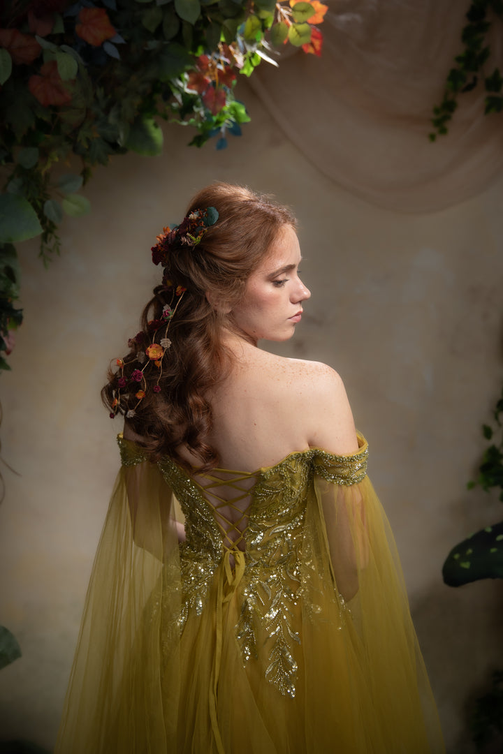 A woman in profile wearing an off-the-shoulder tulle gown with gold lace and embroidery, standing against a soft beige backdrop with floral decorations.