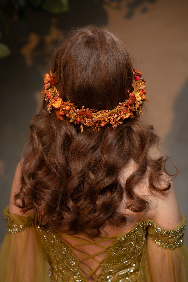 A woman viewed from behind wearing a decorative floral headpiece in warm orange, red, and yellow tones, with long wavy brown hair and an olive-gold dress against a softly blurred brown background.