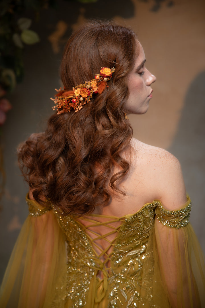 A woman shown from the back wearing a flowing off-the-shoulder gold dress with lace-up back detailing and a floral hair accessory in orange and yellow tones, against a softly lit warm brown background.