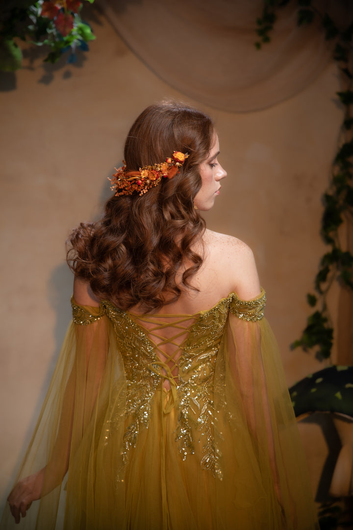 A woman with long, wavy brown hair is shown from the back wearing an off-the-shoulder golden gown with intricate gold embroidery and a lace-up back. She is wearing a small orange floral hair accessory against a warm beige backdrop with decorative foliage.