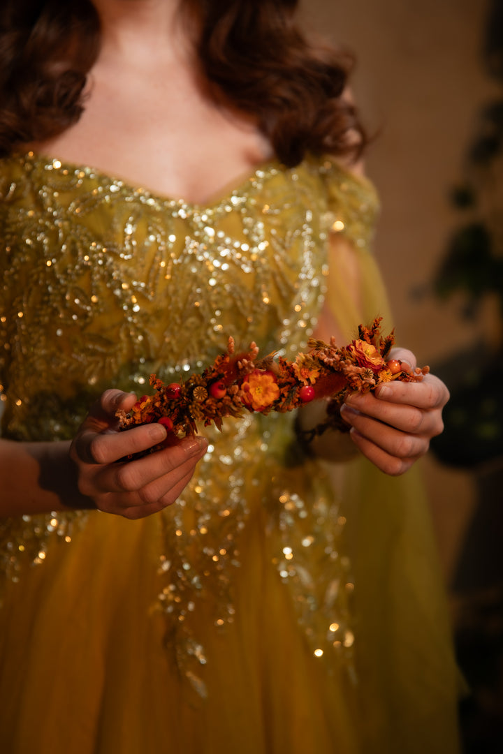 A woman in a sparkling gold sequined gown holds a dried floral crown with orange, red, and brown botanical elements against a warm, softly blurred background.