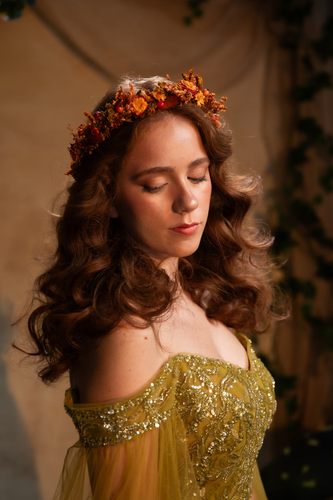 A woman with long wavy brown hair wearing a floral crown and a gold off-the-shoulder embellished gown, captured in warm, moody lighting against a soft beige backdrop.