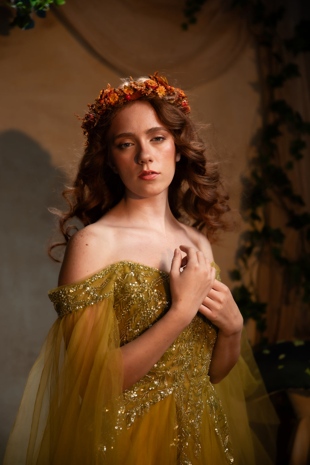 A young woman wearing a sparkling off-the-shoulder yellow gown with gold sequins and a floral crown, posed against a warm, draped studio backdrop with greenery.