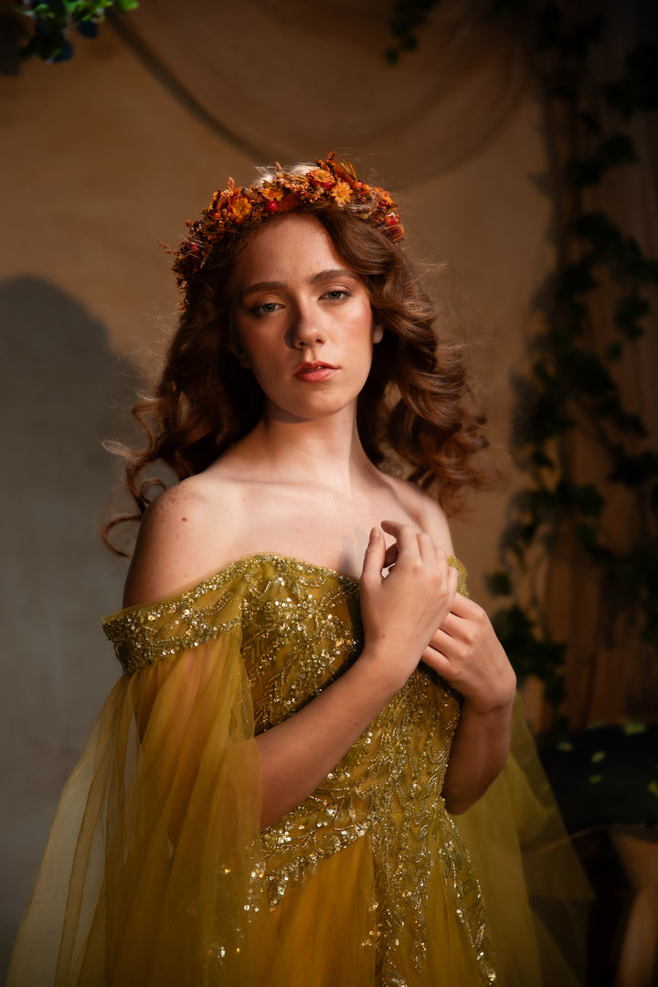 A young woman wearing a sparkling off-the-shoulder yellow gown with gold sequins and a floral crown, posed against a warm, draped studio backdrop with greenery.