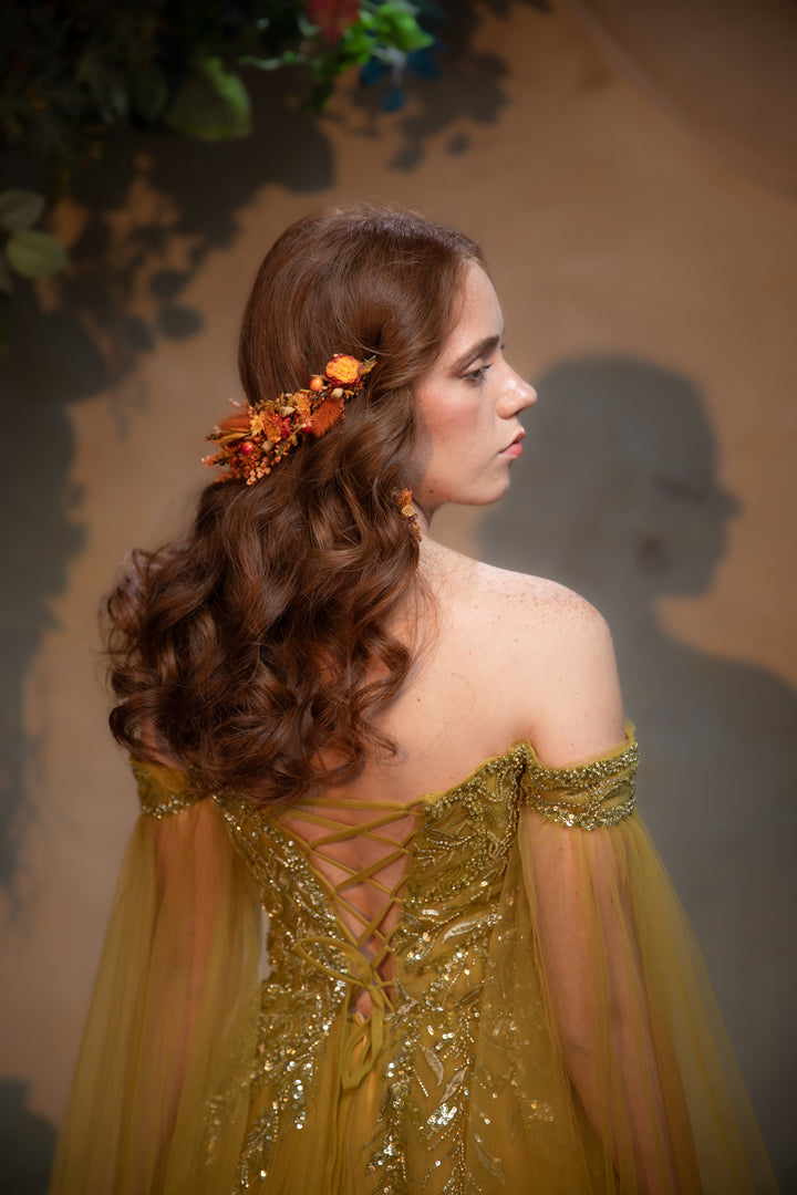 A woman in profile wearing a shimmering off-the-shoulder gold gown with lace-up back details and sheer tulle sleeves, styled with an orange floral hair accessory against a warm beige background.
