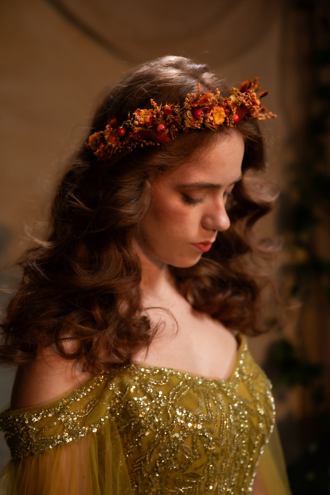 A woman with long wavy brown hair wears an orange and brown floral crown and a gold off-the-shoulder embellished gown, looking down in a dimly lit setting.