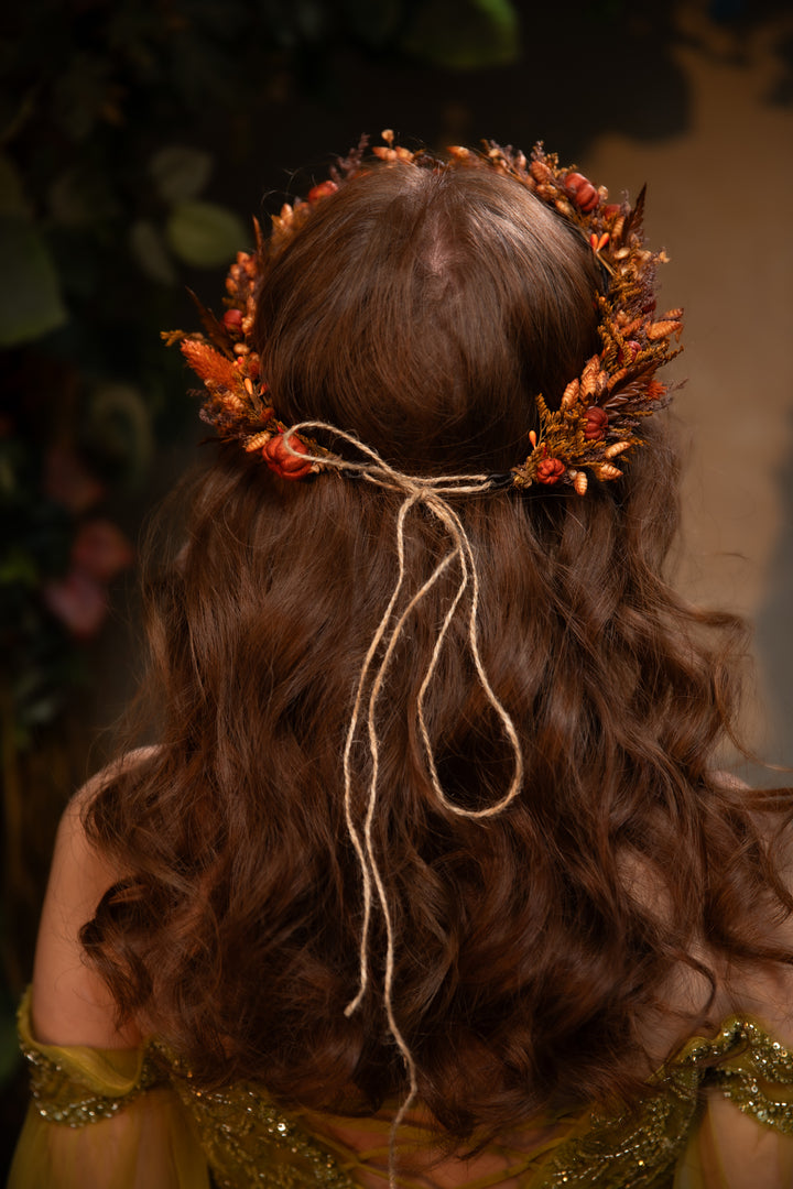 A woman viewed from behind wearing a decorative floral hair crown with orange and brown leaves tied with a twine bow. She has long, wavy brown hair and is wearing a gold embellished dress against a dark, blurred background.