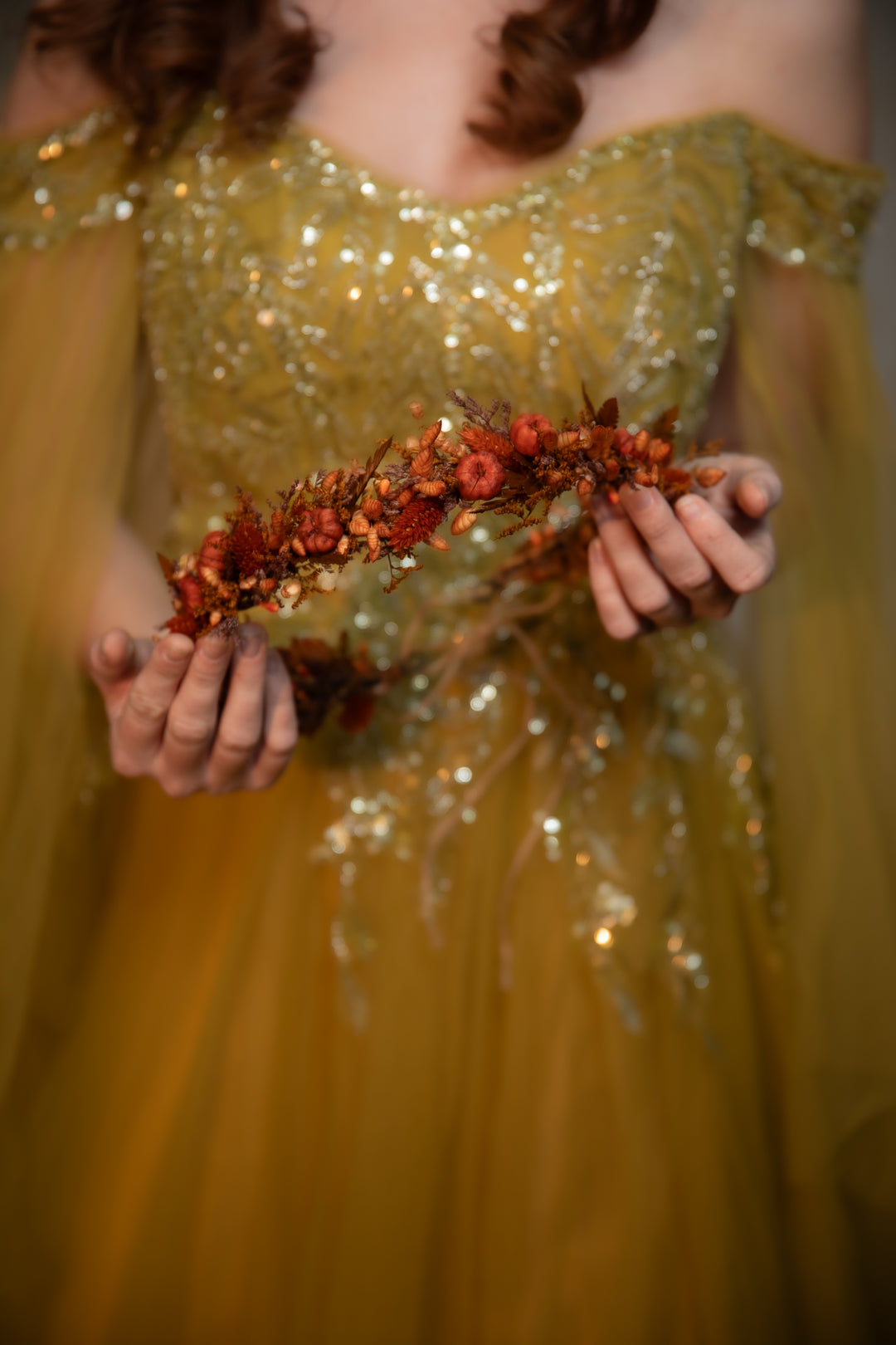 A close-up of a person wearing a shimmering off-the-shoulder gown and holding a dried floral crown in both hands.