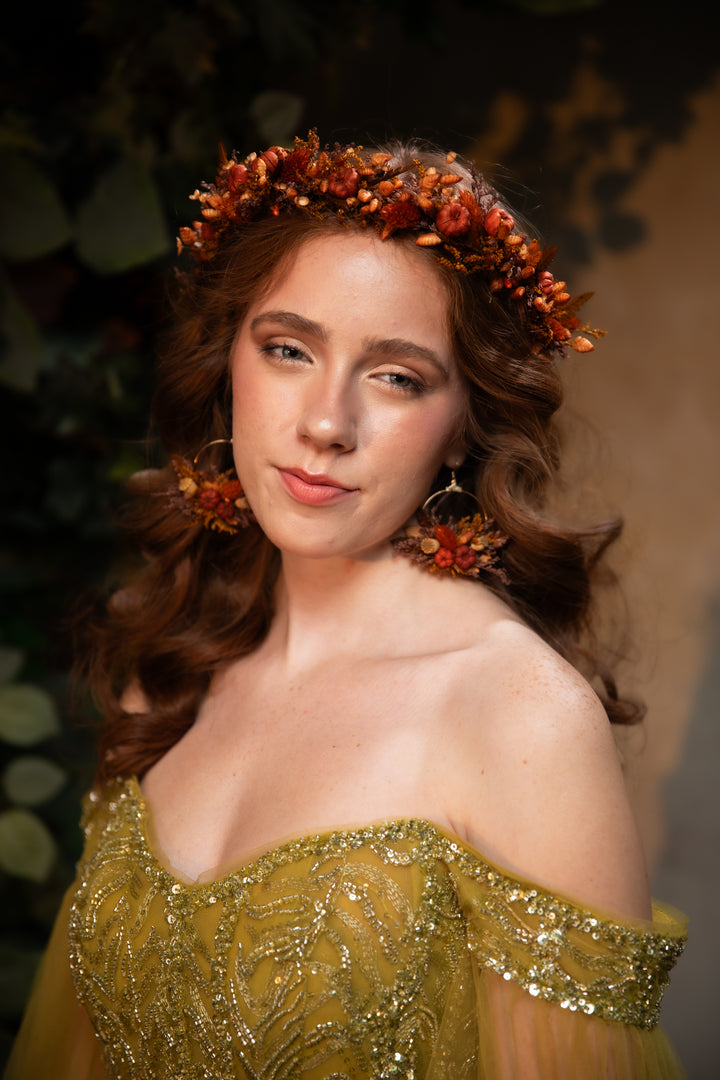 A woman wearing a floral crown with orange and brown flowers, matching dangling earrings, and an off-the-shoulder olive green sequined gown, photographed against a dark, softly blurred background.