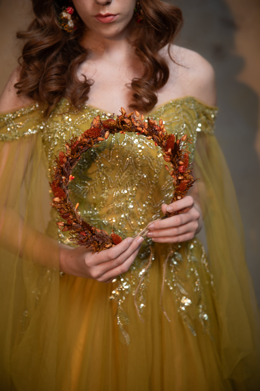 A woman in an off-the-shoulder sequined gown holds a circular wreath made of metallic gold and rust-orange leaves.