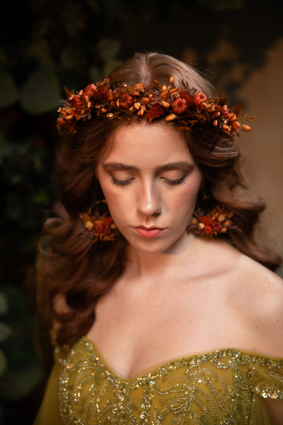 A woman with long wavy brown hair wearing a floral crown in rust orange and brown tones, paired with matching floral earrings and a gold embellished off-the-shoulder dress.