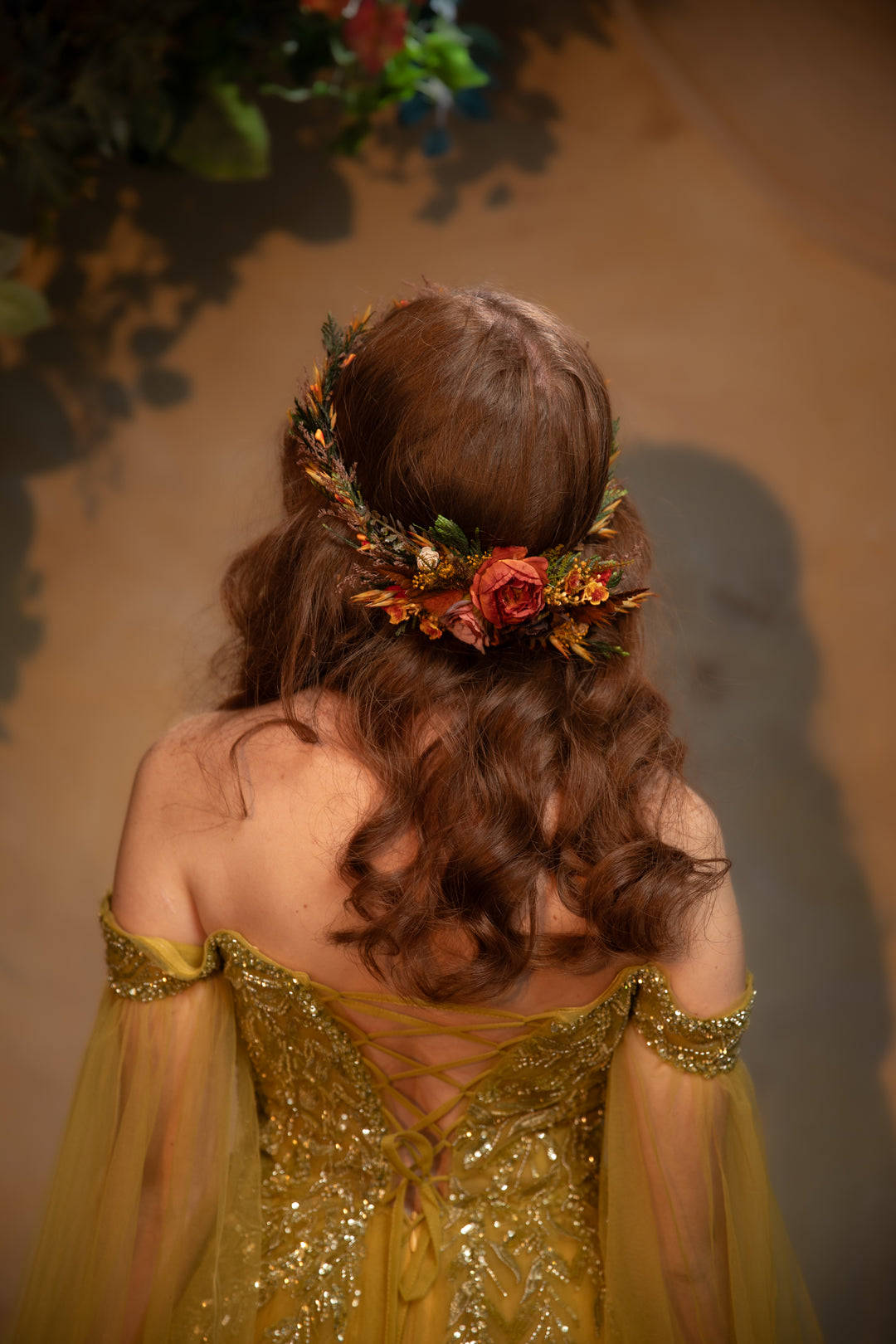 A woman viewed from behind wearing a flowing gown with sheer draped sleeves and a floral crown with orange and ochre flowers, set against a warm brown background.