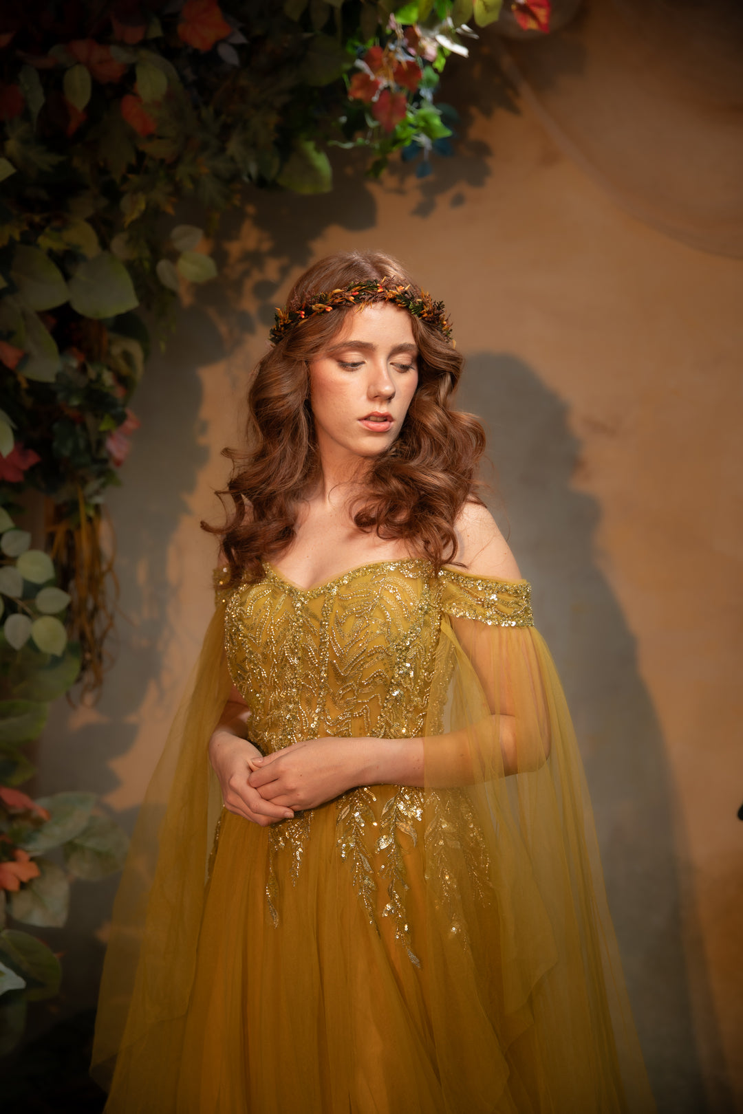 A woman wearing a off-the-shoulder gown with sequined embellishments and a sheer tulle cape, styled with a floral crown, standing in front of a warm-toned backdrop with floral decorations.
