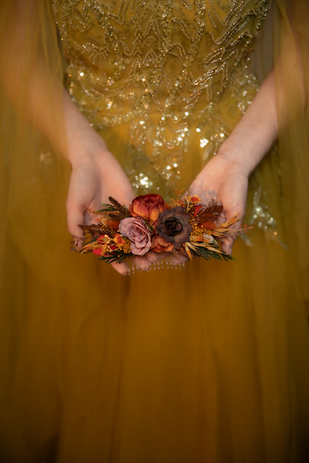 A close-up of a person wearing a shimmering embellished dress and holding a small floral hair accessory with muted orange, burgundy, and mauve flowers.