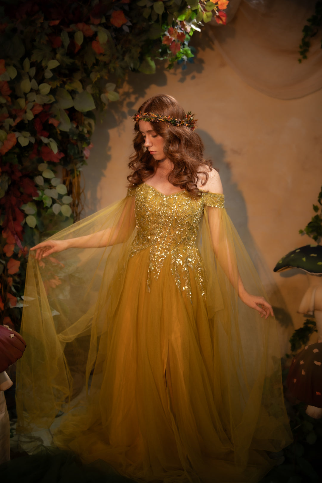 A woman wearing a flowing off-the-shoulder gown with heavy gold beadwork and a sheer draped overlay, posed in front of a warm-toned backdrop with decorative foliage and flowers.