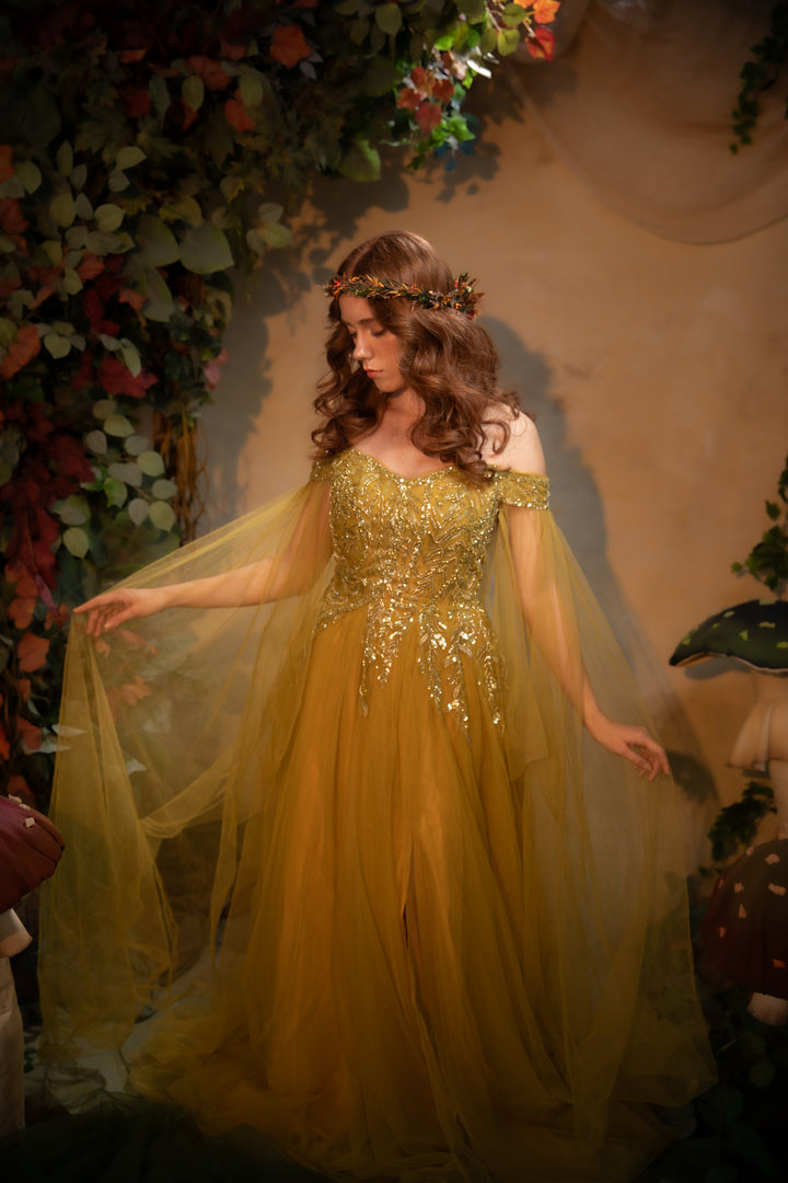 A woman wearing a flowing off-the-shoulder gown with heavy gold beadwork and a sheer draped overlay, posed in front of a warm-toned backdrop with decorative foliage and flowers.