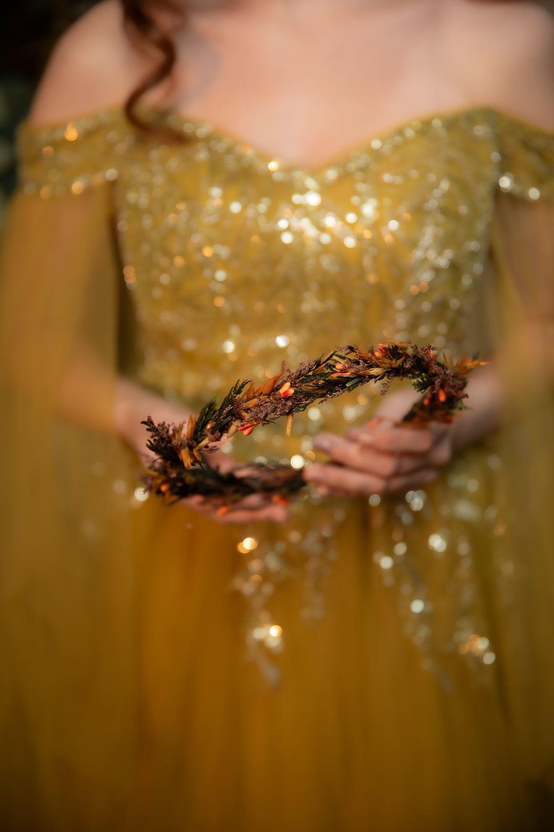 A woman in a sparkling off-the-shoulder gown holds a dark brown pine wreath with small orange berries in her hands.