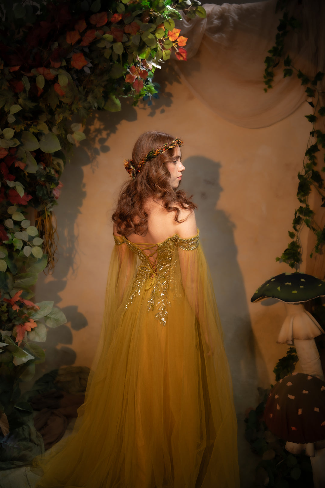 A woman wearing a flowing tulle gown with gold embellishments, photographed in a fantasy-style forest scene with foliage, mushrooms, and warm ambient lighting.
