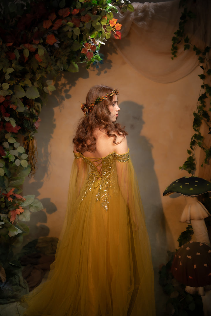 A woman wearing a flowing tulle gown with gold embellishments, photographed in a fantasy-style forest scene with foliage, mushrooms, and warm ambient lighting.