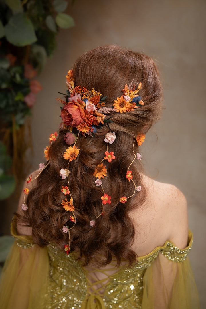 A woman shown from the back wearing a styled brown wavy hairstyle adorned with orange and yellow floral hair accessories, a small pink bow, and delicate vine details. She is wearing a gold off-the-shoulder embellished dress against a soft-focus background with green foliage.
