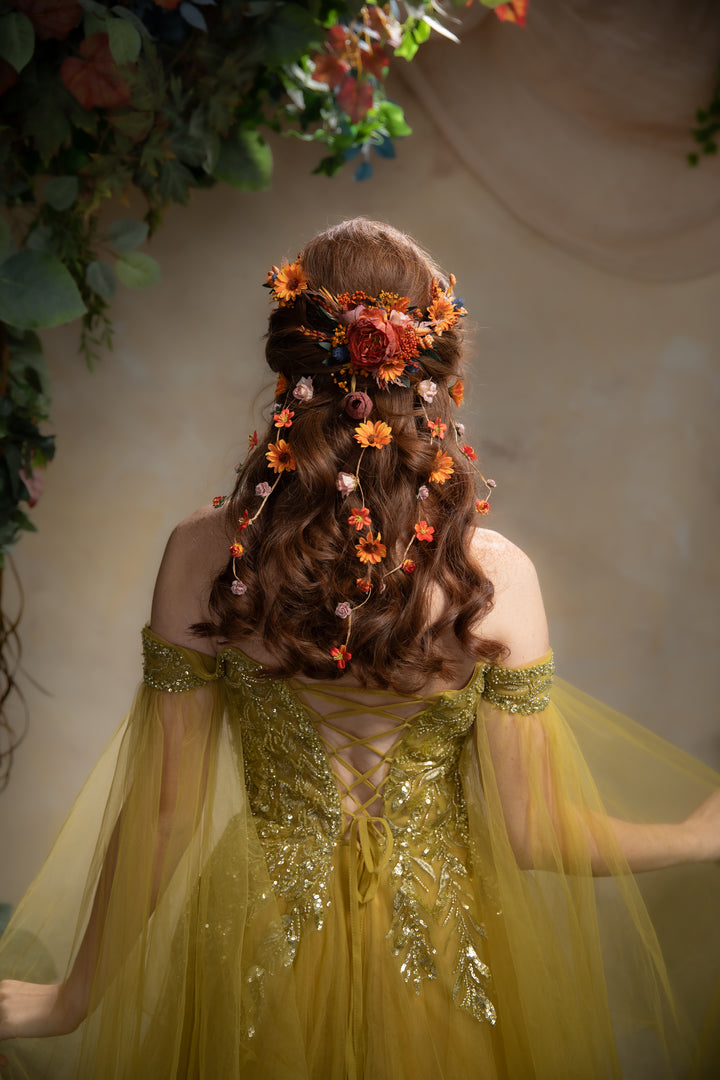 A woman viewed from behind wearing a flowing yellow gown with sheer tulle sleeves and ornate gold embellishment, styled with a brown wavy hairstyle adorned with orange and red flowers. The background is a soft beige wall with hanging green foliage.