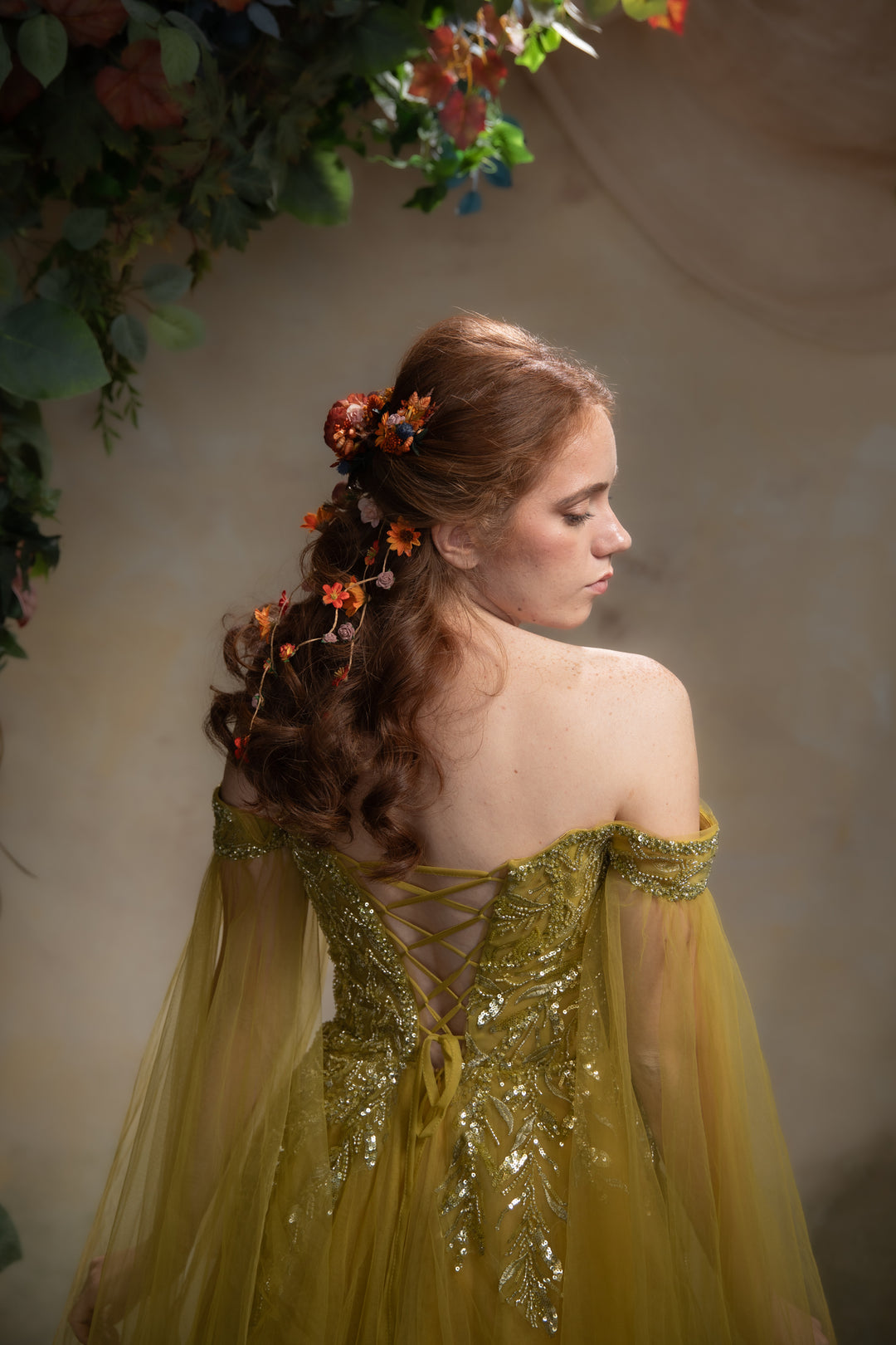 A woman in profile wearing an off-the-shoulder yellow gown with lace-up back detailing and sheer draped sleeves, styled with orange flowers in her hair against a soft beige backdrop with floral decorations.