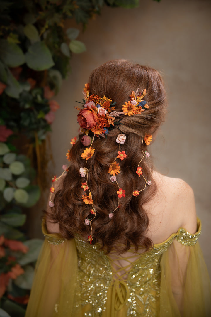 A woman shown from behind wearing a decorative floral hairpiece with orange, peach, and pink flowers and green leaves woven into her styled brown hair. She is wearing a gold off-the-shoulder sequined dress, set against a soft-focus background of green foliage and a neutral wall.