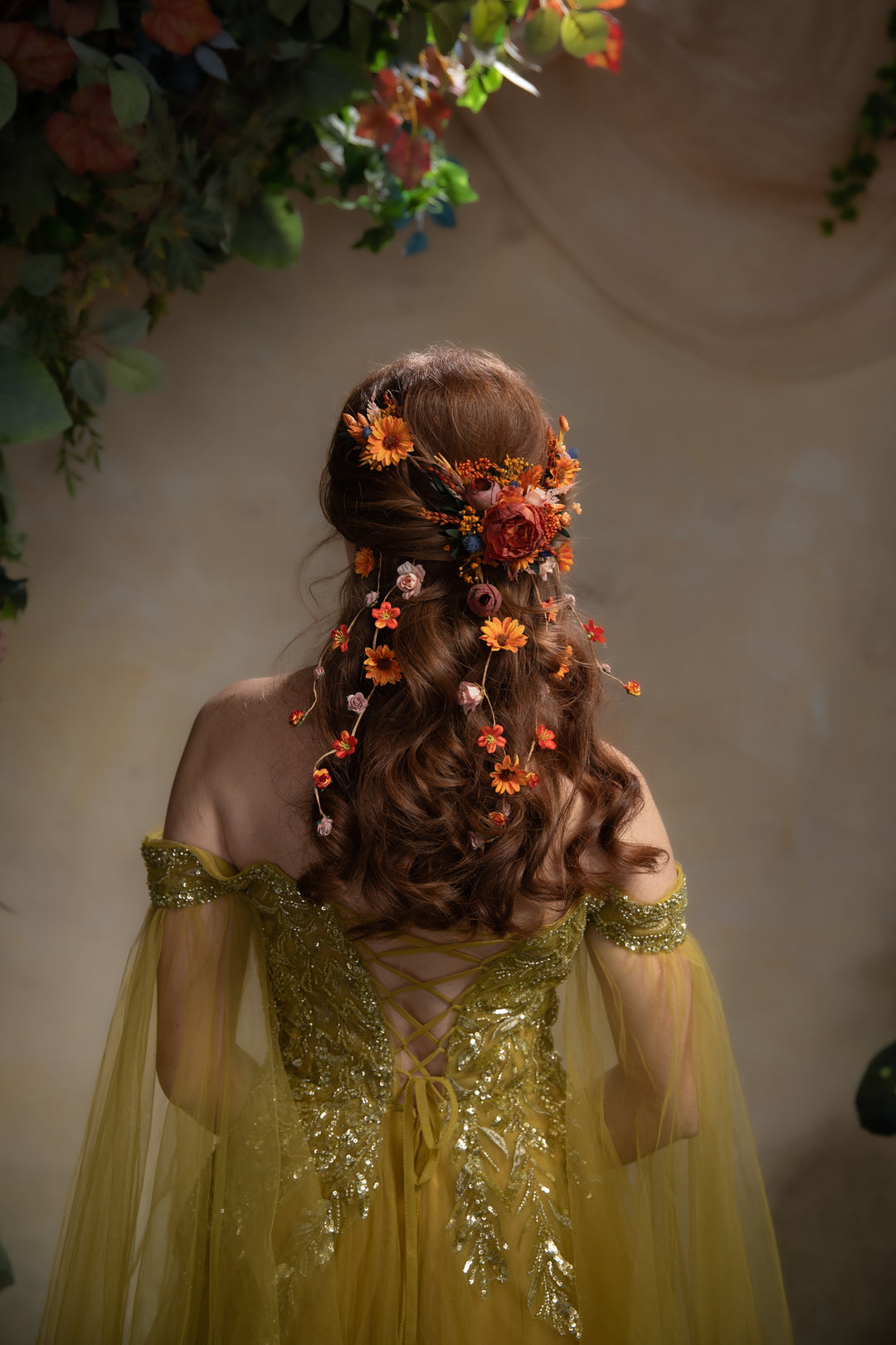 A woman viewed from behind wearing a flowing yellow gown with gold embellishment and a styled brown updo decorated with orange and pink flowers. She stands against a warm beige wall with floral greenery hanging at the top.