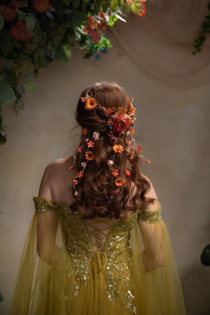 A woman viewed from behind wearing a flowing yellow gown with gold embellishment and a styled brown updo decorated with orange and pink flowers. She stands against a warm beige wall with floral greenery hanging at the top.
