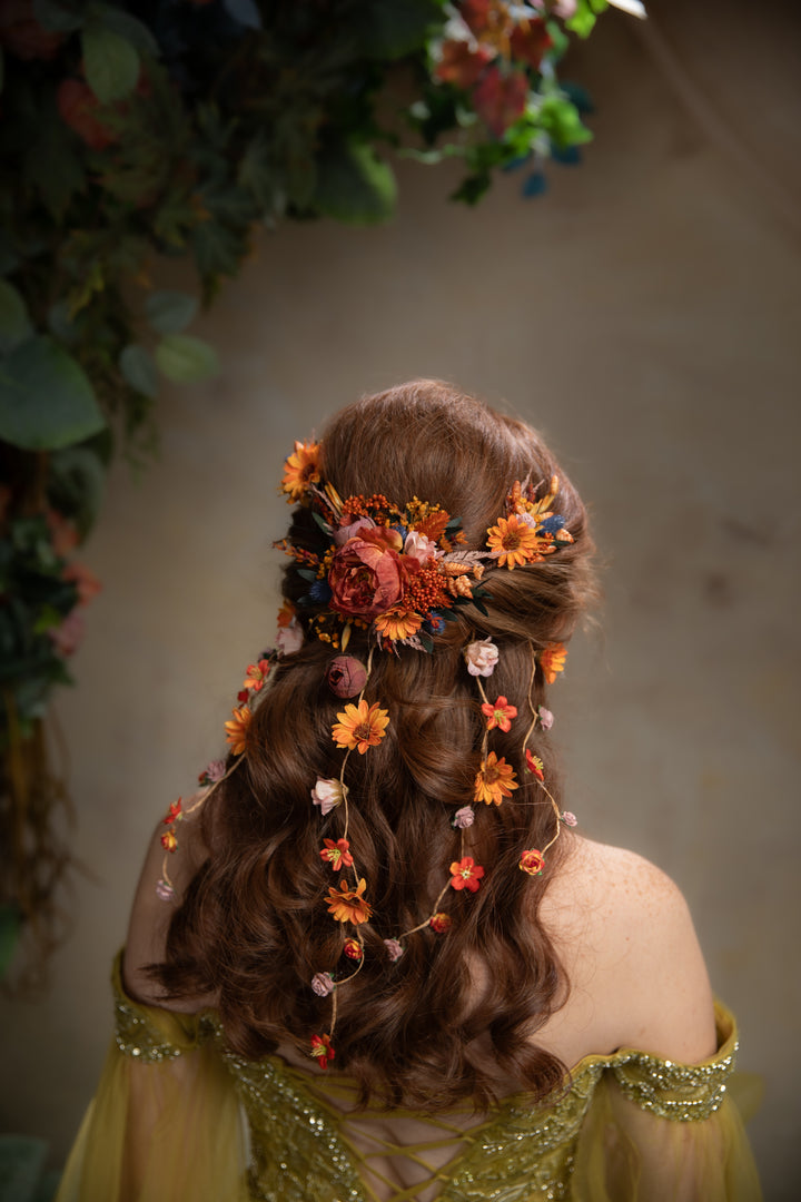 A woman shown from behind wearing a decorative floral hairpiece with orange, peach, and pink flowers and greenery, styled in a wavy brown updo with loose curls. She wears a pale yellow off-the-shoulder dress against a softly blurred neutral background with hanging foliage.