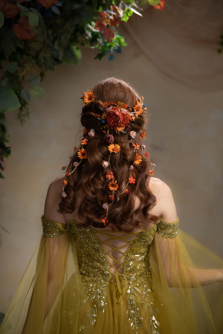A woman viewed from behind wearing a flowing yellow gown with lace-up back details and sheer tulle sleeves, styled with a floral hairpiece made of orange and red flowers and green leaves against a soft beige backdrop with hanging foliage.