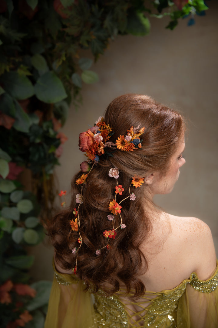A woman shown from the back wearing a styled brown wavy hairstyle adorned with orange, yellow, and pink dried flowers and small vine accents. She is wearing a gold off-the-shoulder dress, set against a soft beige background with green foliage on the left.