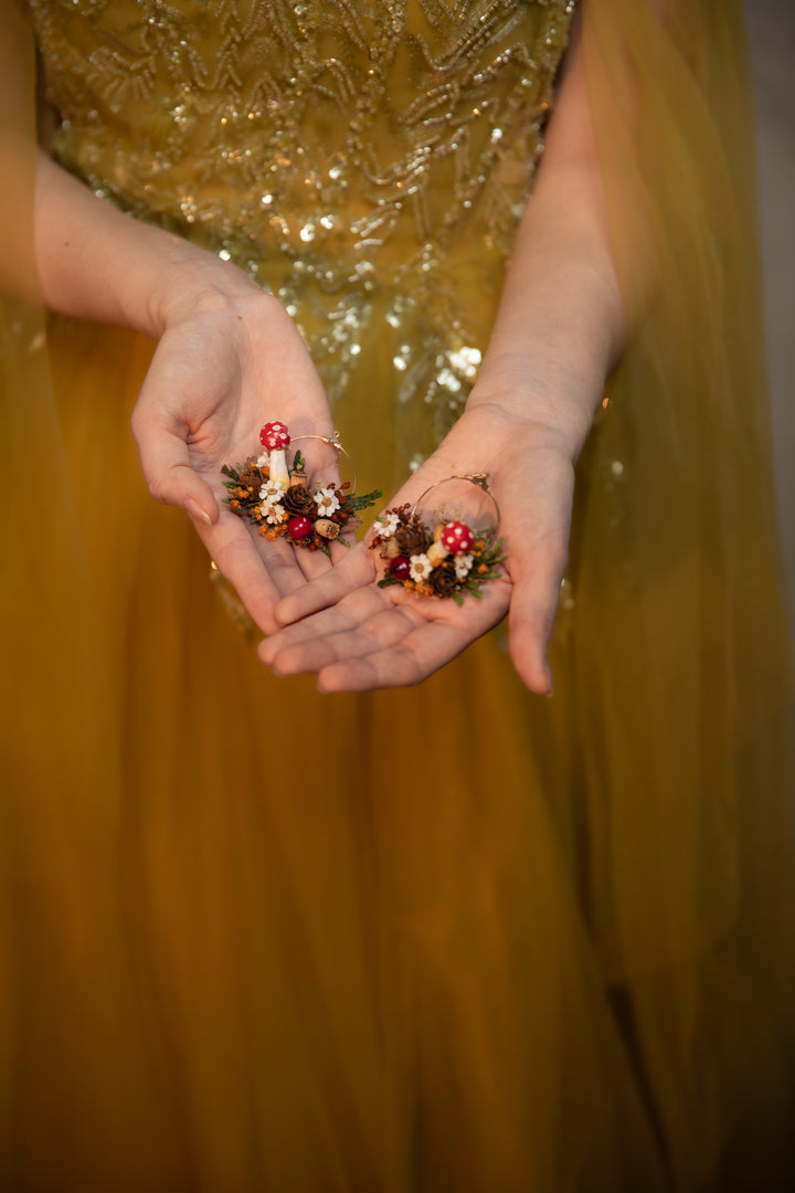 A close-up of a person in a gold sequined dress holding two searrings with mushrooms, cream flowers, and green leaves in their hands.