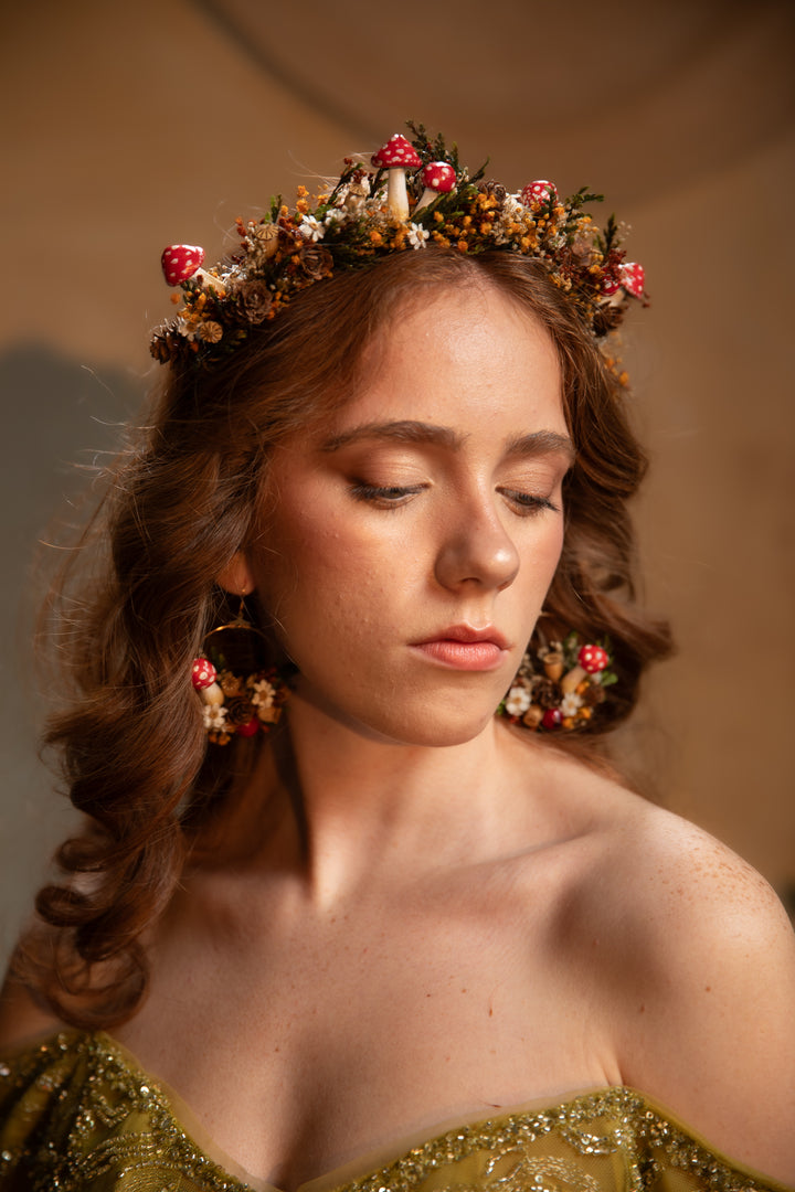 A woman with closed eyes wearing a floral crown and matching dangling earrings, photographed in warm studio lighting against a soft beige background.