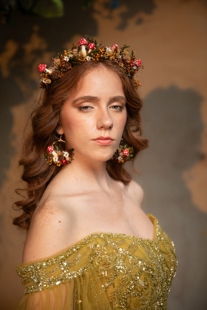 A woman with wavy brown hair wearing a floral crown and matching dangling earrings, posed in an off-the-shoulder gold embellished gown against a warm brown studio background.
