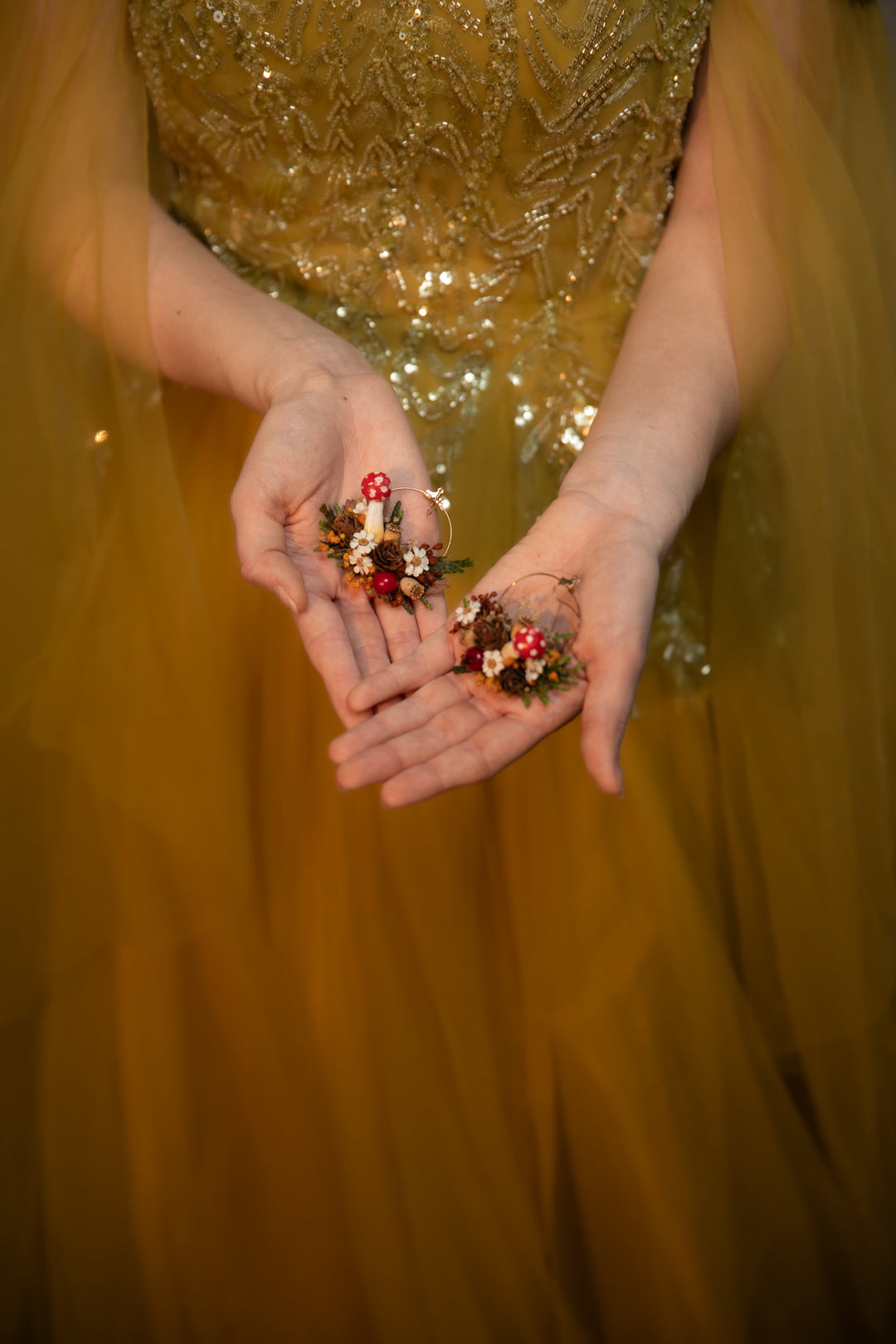 A close-up of a person in a shimmering gold sequined dress holding circle dangle earrings  in their hands against a soft, out-of-focus gold background.