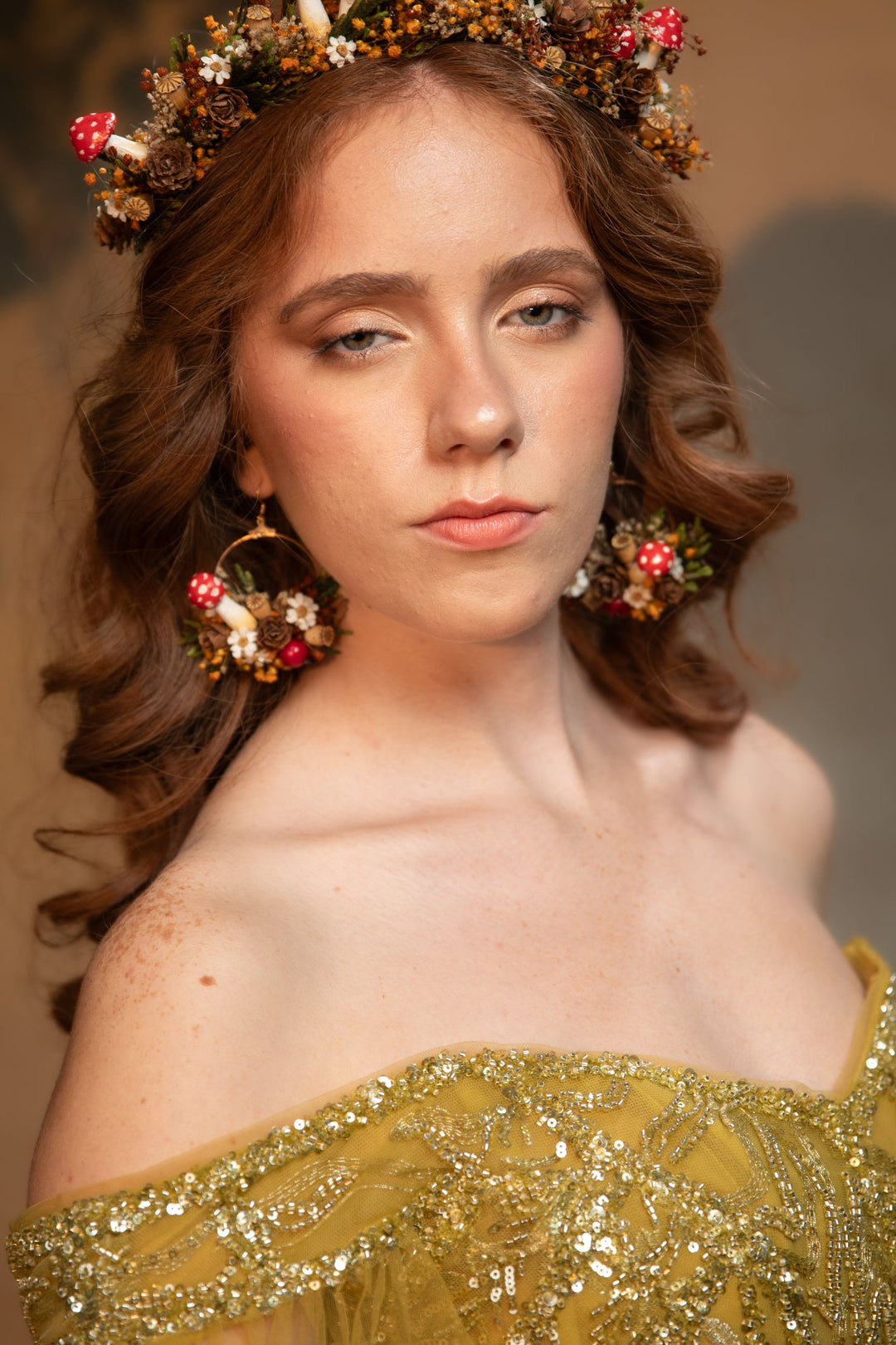 A woman wearing a floral crown with red mushrooms, matching floral earrings, and an off-the-shoulder gold embellished gown, photographed against a warm brown studio backdrop.