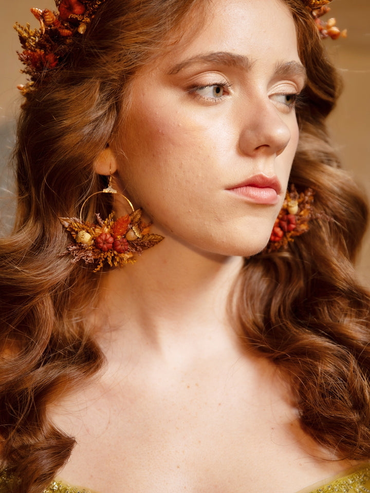 A close-up portrait of a woman with long wavy brown hair wearing large, ornate gold hoop earrings adorned with dried orange and earthy botanical elements, along with a matching floral headpiece.